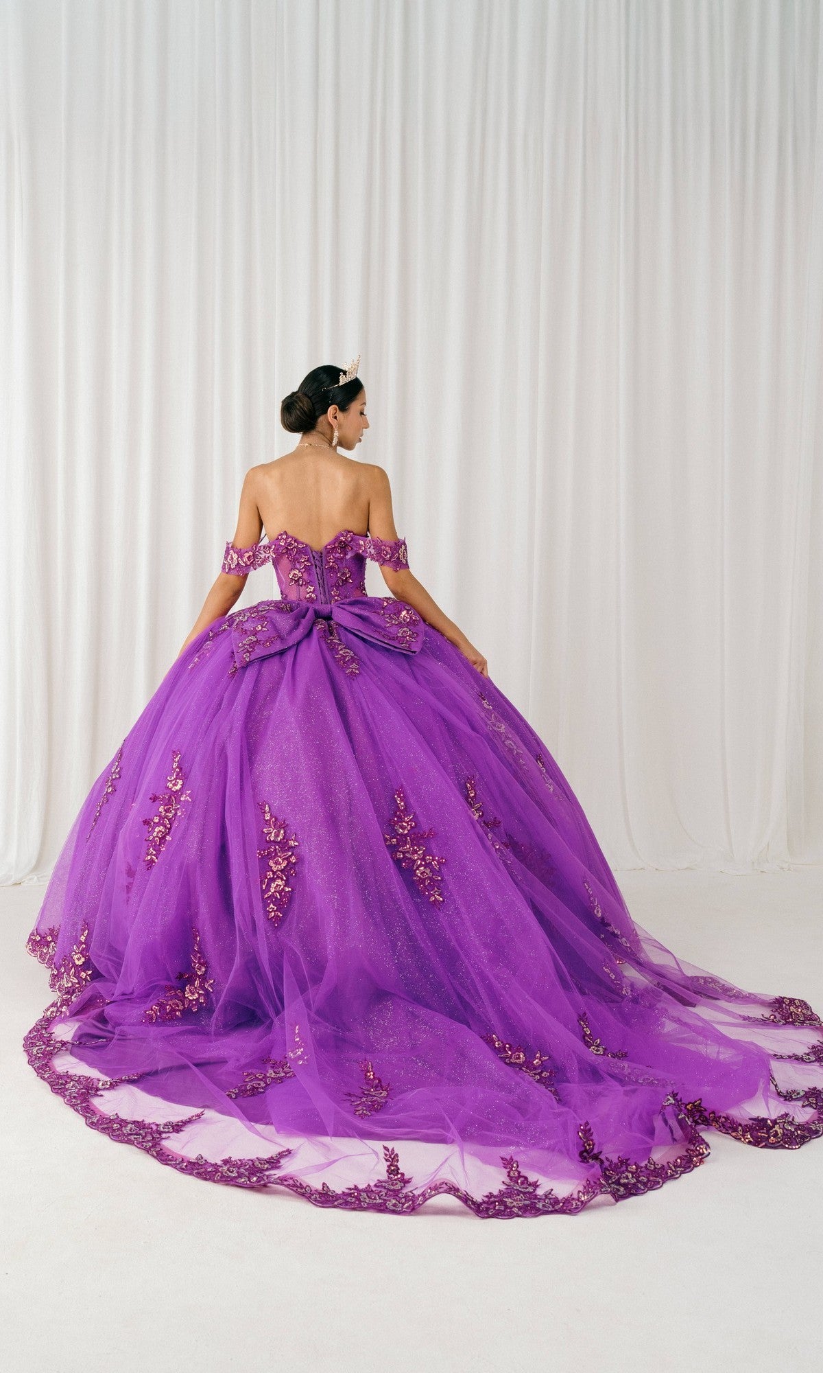 A woman wears the Quinceanera Dress 8405JQ by Cinderella Couture, featuring an off-the-shoulder ball gown design with floral embellishments and a large bow, standing against a white draped backdrop.